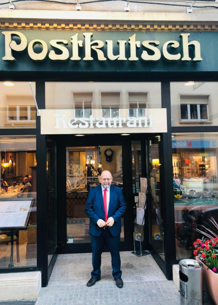 Man in suit outside Postkutsch restaurant entrance.