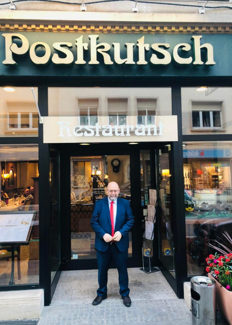 Man in suit outside Postkutsch restaurant entrance.