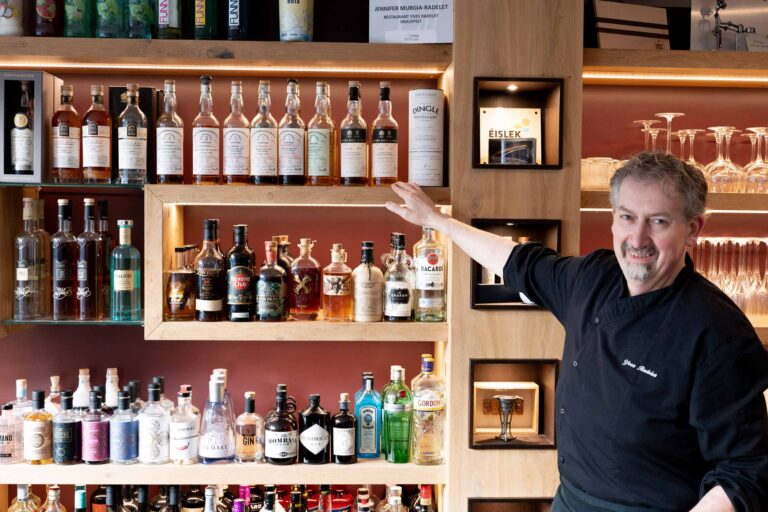 Smiling bartender showcasing liquor bottles on shelves.