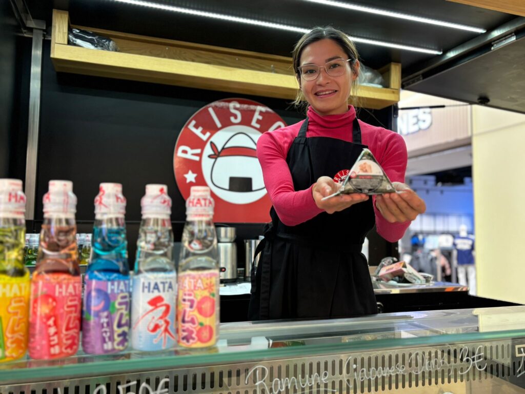 Woman presents onigiri and flavored drinks at food stall.