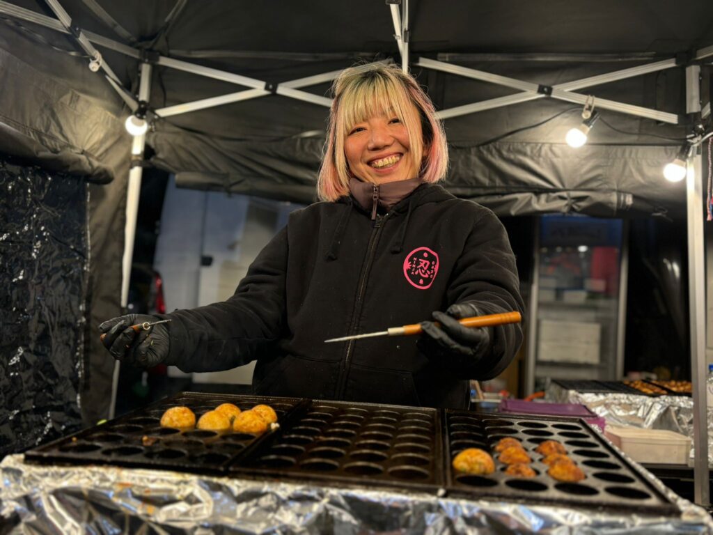 Chef smiling, cooking takoyaki at food stall.